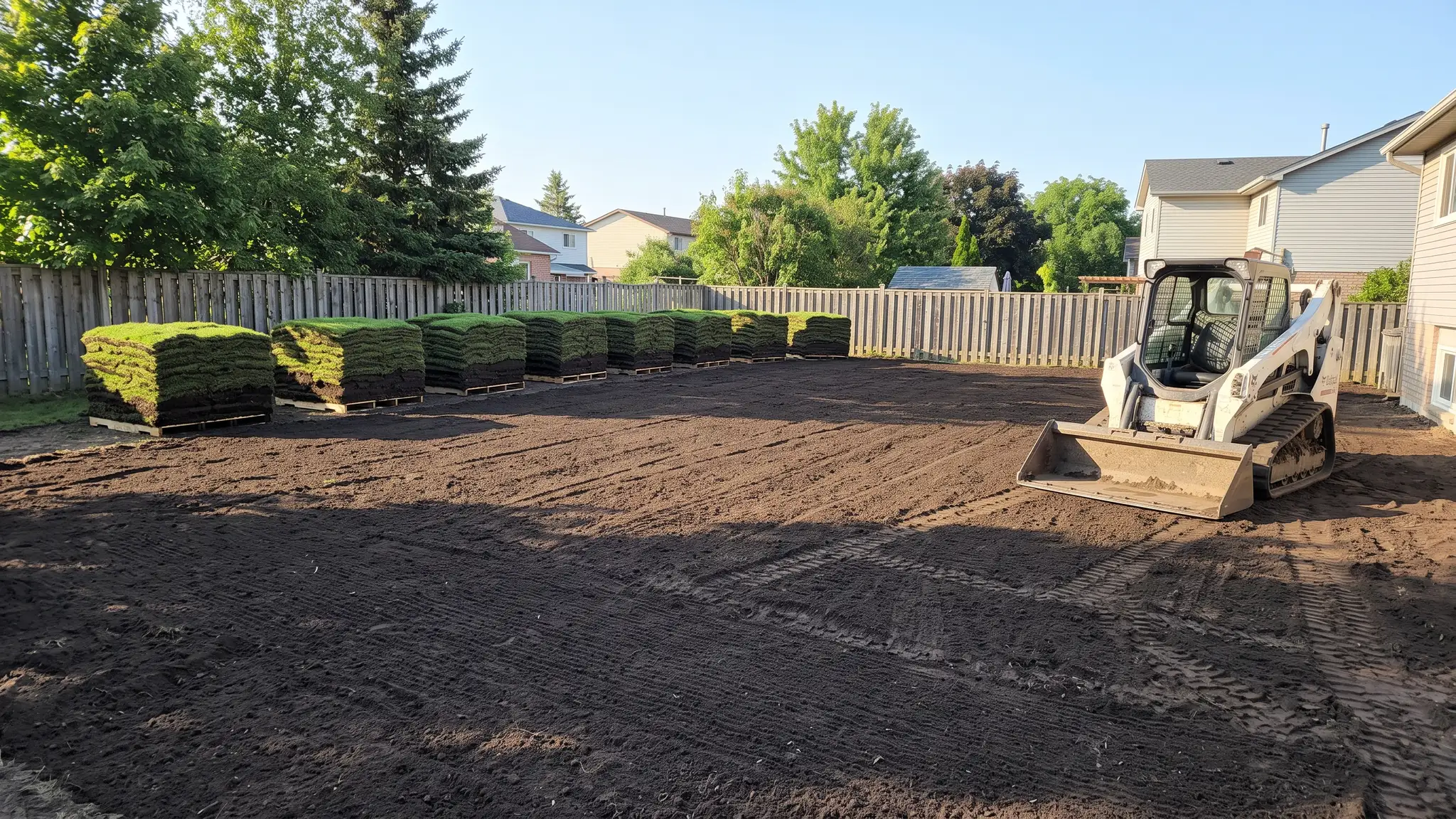 A freshly graded, raked dark soil yard ready for sod with pallets of green sod at the edge and a compact skid steer parked nearby