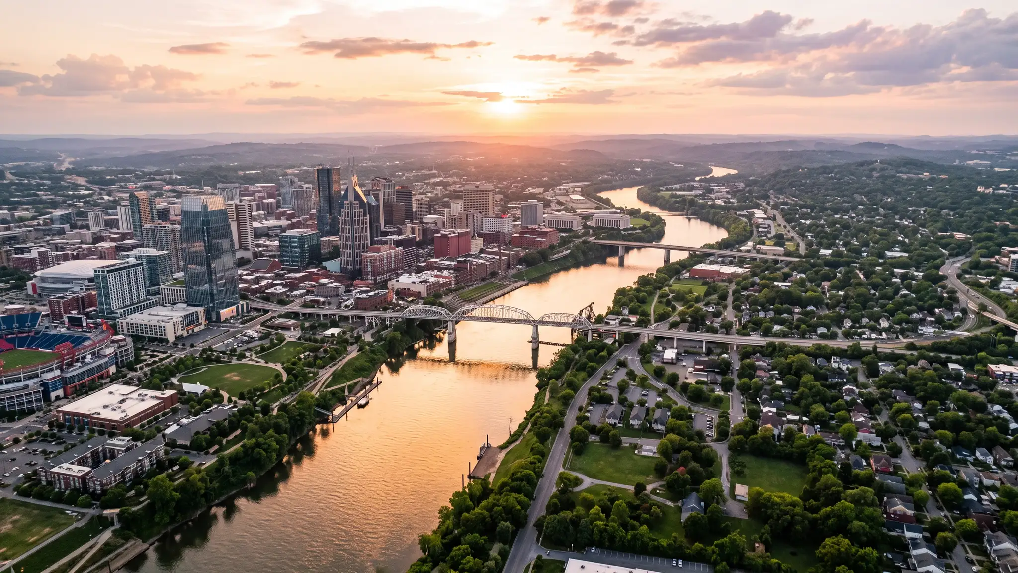 Aerial view of the Cumberland River winding past the Nashville skyline and green residential neighborhoods at sunset with golden light on the water